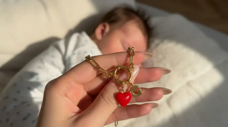 Main de femme qui tient un bijou souvenir connecté Joylink avec un charm coeur rouge et une initiale G, dans lequel sont conservés des souvenirs de naissance du bébé en arrière plan